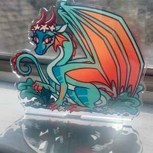 May include: A teal and orange dragon figurine with a white flower crown, sitting on a clear acrylic base. The dragon has a curled tail and is surrounded by white flowers.