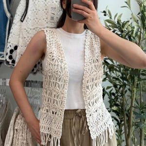 Può includere: Un gilet senza maniche all'uncinetto color bianco sporco caratterizzato da un intricato motivo floreale su tutto il corpo. Il capo presenta un design aperto sul davanti ed è rifinito con una fila di lunghe frange pendenti lungo l'orlo inferiore.