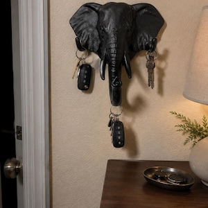 May include: A black wall-mounted elephant head key holder featuring three hooks for hanging items. The hooks are positioned at the elephant's ears and the tip of the trunk. Several sets of metal keys and black key fobs are hanging from the hooks.
