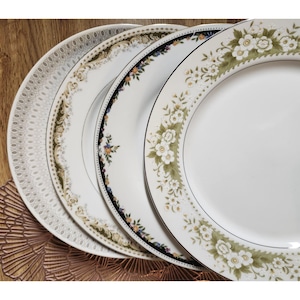 May include: A set of four round ceramic dinner plates stacked together. Each plate features a distinct rim pattern, including white floral motifs with green leaves, a dark blue border with small flowers, gold scrollwork, and a grey geometric design.