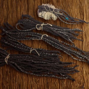 Puede incluir: Varios paquetes de rastas de color gris oscuro con costuras blancas se disponen sobre una superficie de madera. Cada paquete está atado con hilo. También hay un paquete más pequeño de rastas, con envoltorio plateado y detalles azules.