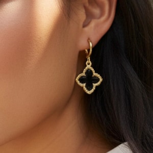 May include: A gold hoop earring featuring a dangling four-leaf clover charm. The charm has a black center inlay surrounded by a textured gold border. The earring is secured with a small gold hoop that attaches to the clover-shaped pendant.