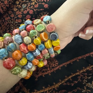 May include: A multi-strand bracelet featuring numerous small, round, and irregularly shaped ceramic beads in a vibrant array of colors including red, blue, yellow, green, orange, and pink, with some beads displaying a speckled or textured finish.