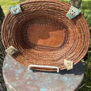 May include: Oval-shaped woven basket with a wooden base and decorative corner accents. The basket is made of natural-colored reeds and features four decorative corner pieces with a button-like design. A white metal handle is attached to the base.