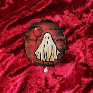 May include: A white ghost with a red balloon painted on a wooden round slice. The background is red.