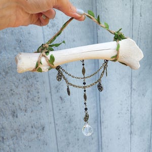 May include: A white bone wall hanging suspended by a twine rope adorned with green fabric leaves. Attached to the bone are multiple antique bronze-colored metal chains featuring small pentagram charms, black beads, and a clear faceted crystal drop.