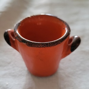 May include: Small, orange ceramic cup with two small handles. The rim and handles have a dark brown, speckled trim. The cup is sitting on a white, textured surface. The cup is likely vintage or handmade.