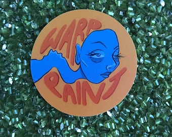 May include: A round sticker with the words "WARP PAINT" in orange lettering. The sticker features a blue illustration of a face with closed eyes and a flowing shape. The background is a bed of green beads.