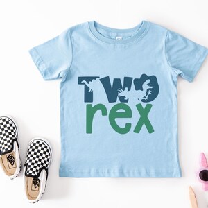 May include: A light blue toddler t-shirt with the words "TWO rex" in dark blue and green. The design includes dinosaur silhouettes. Black and white checkered slip-on shoes are next to the shirt.