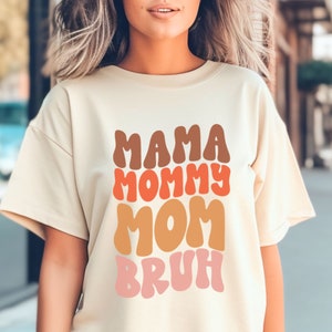 以下が含まれることがあります： オレンジ、ブラウン、ピンクのレトロなグラフィックが施された薄いベージュのTシャツ。Tシャツには「ママ、ママ、ママ、ブル」と書いてあります。