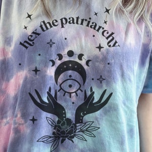 Puede incluir: Una camiseta tie-dye con un estampado negro de una luna creciente, estrellas y manos con el texto "hex the patriarchy" sobre el diseño.