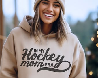 May include: Beige hoodie with the text "IN MY Hockey mom ERA" in a black script font. The hoodie has a drawstring hood and a kangaroo pocket. The image shows a person wearing the hoodie.