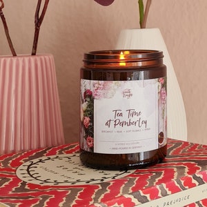 May include: A brown glass candle with a white label that reads "Tea Time at Pemberley". The candle is sitting on top of two books, one with a red and white patterned cover and the other with a cream and red patterned cover. The books are titled "Pride and Prejudice" and "Persuasion" by Jane Austen.