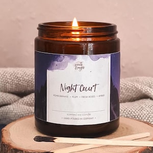 Könnte beinhalten: Eine brennende Sojakerze in einem bernsteinfarbenen Glas mit einem Etikett, das Night Court, Pomegranate, Plum, Fresh Roses, Amber und Hand-Poured in Germany zeigt. Zwei Holzstreichhölzer mit verkohlten Enden liegen auf einer runden Holzscheibe.