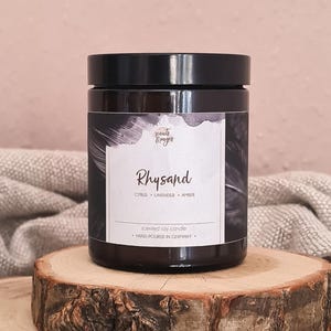 Op de afbeelding: Een kaars in een donker amberkleurige glazen pot met een zwart deksel en een wit etiket. Het etiket bevat de tekst Scents & Pages, Rhysand, Citrus, Lavender, Amber en Hand-Poured in Germany. De kaars staat op een ronde houten schijf.