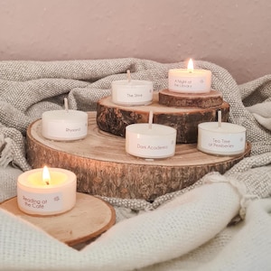 Puede incluir: Cinco velas de té blancas con diferentes nombres con temática de libros sobre una superficie de madera. Las velas están encendidas y los nombres son "Reading at the Cafe", "The Shire", "A Night at the Library", "Rhysand", "Dark Academia" y "Tea Time at Pemberley".