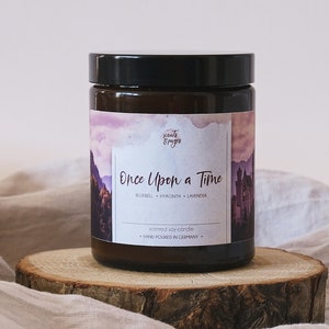 Könnte beinhalten: Eine braune Glaskerze in einem Glas mit schwarzem Deckel. Das Etikett zeigt eine Aquarell-Illustration einer Burg und Berge mit einem rosa und violetten Himmel. Der Text auf dem Etikett lautet "scents & pages", "Once Upon a Time", "Bluebell, Hyacinth, Lavender", "Scented Soy Candle", und "Hand Poured in Germany."