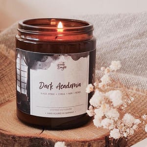 Könnte beinhalten: Eine Duftkerze aus Sojawachs in einem braunen Glasgefäß mit einem Etikett, das Dark Academia, Black Amber, Citrus, Rum, Roses und Hand Poured in Germany zeigt. Die Kerze hat einen brennenden Docht und steht auf einer Holzscheibe neben Trockenblumen.