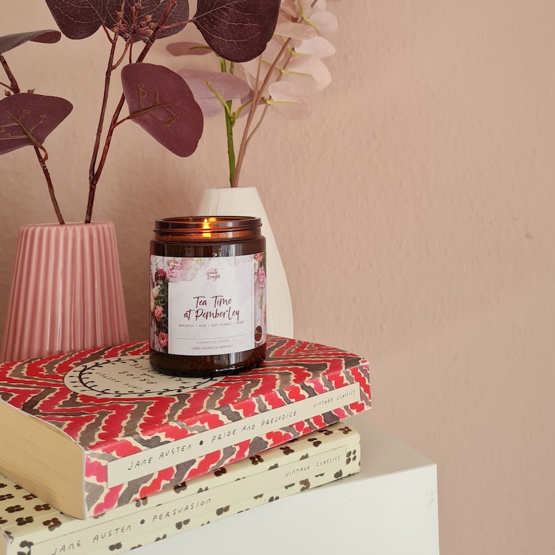May include: A brown glass candle with a white label that reads "Tea Time at Pemberley". The candle is sitting on top of two books, one with a red and white patterned cover and the other with a cream and red patterned cover. The books are titled "Pride and Prejudice" and "Persuasion" by Jane Austen.
