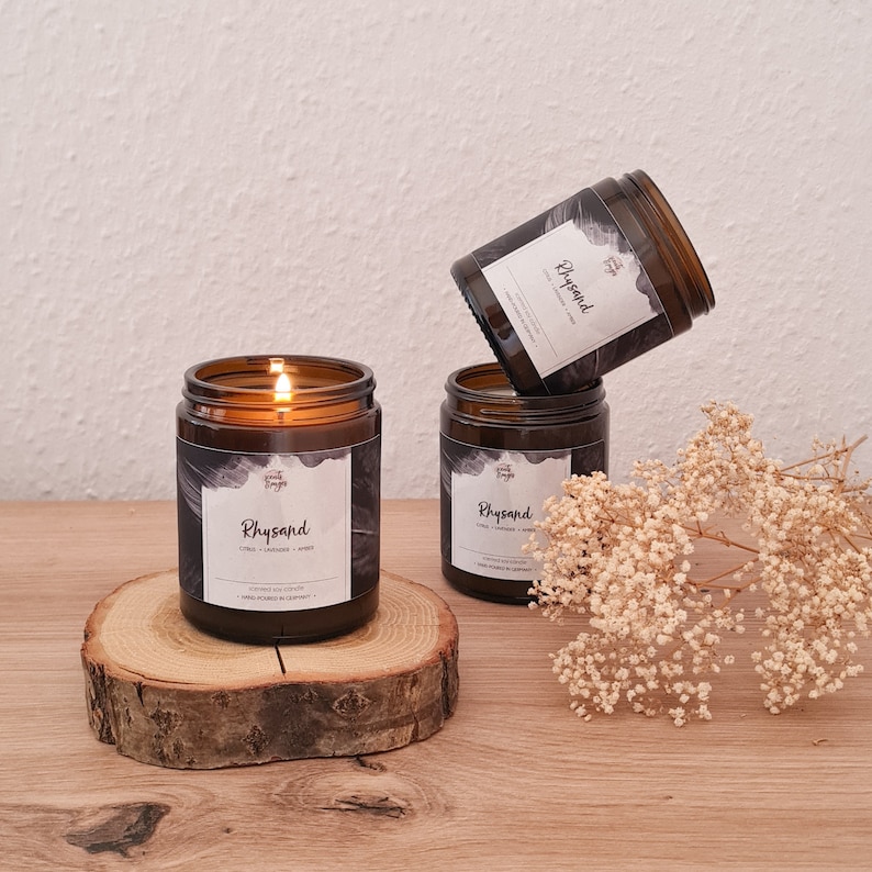 A lit candle called Rhysand rests on a wooden coaster. Behind it, to its right, there are two more of the same candle, one on top of other. The candle comes in an amber jar, and its label is mainly black with a gray feather slightly visible.