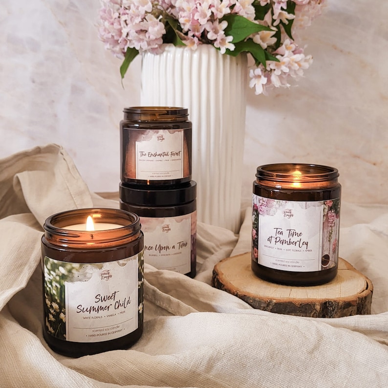 May include: Four amber glass candle jars with labels and floral designs. The candles are lit and have a warm glow. The labels read "The Enchanted Forest", "Once Upon a Time", "Sweet Summer Child", and "Tea Time at Pemberley".