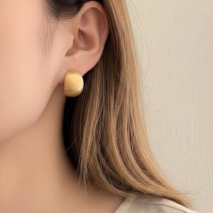 Pode incluir: Um close-up de um brinco de botão quadrado arredondado dourado. O brinco tem um acabamento fosco sólido e é usado na orelha. O fundo é de cor bege neutra.