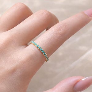May include: A gold-colored ring with a band of small, turquoise-colored stones. The ring is worn on a finger, showcasing the delicate design and vibrant color of the gemstones. The ring is a piece of jewelry.