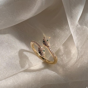 May include: A gold-colored snake ring with a wrap-around design. The snake's head and body are adorned with small, clear and green gemstones. The ring is set against a white, textured background.
