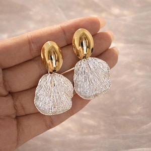 Könnte beinhalten: Ein Paar gold- und silberfarbene Ohrringe. Jeder Ohrring hat einen ovalen, goldenen oberen Teil und einen strukturierten, silberfarbenen, blütenblattförmigen unteren Teil. Die Ohrringe sind zum Hängen konzipiert.