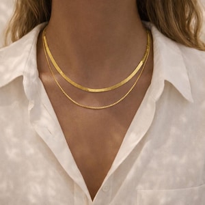 Puede incluir: Dos collares dorados superpuestos. El collar superior es una cadena de espiga plana y ancha, y el inferior es una delicada cadena de caja. Los collares se usan sobre una camisa blanca abotonada.