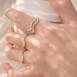 May include: Two rings, one silver and one gold, are displayed on a hand. The silver ring has a wavy design, while the gold ring has a similar, but simpler, wavy pattern. The rings are likely made of metal and are designed to be worn as jewelry.