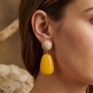 Puede incluir: Un par de pendientes. Cada pendiente presenta un perno esférico de color beige claro y un colgante grande, en forma de lágrima, de color amarillo brillante. Los pendientes cuelgan de un metal dorado. Los pendientes se llevan en la oreja de una persona.