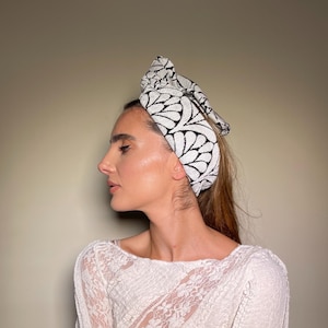 Op de afbeelding: Een witte haarband met een zwart bladerpatroon wordt op het hoofd gedragen. De haarband is bovenaan met een strik vastgebonden. De persoon draagt een witte kanten top met lange mouwen en een ronde hals. De achtergrond is effen en neutraal.