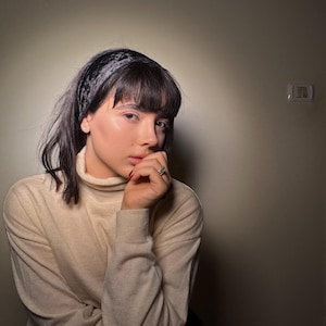 May include: A person wearing a beige turtleneck sweater and a black patterned headband. The person is wearing a ring on their finger. The background is a solid, neutral color.