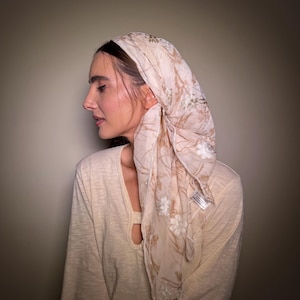May include: A light beige scarf with a floral design, worn over the head and shoulders. The scarf has white and gold-coloured flowers and leaves. The person is wearing a light beige long-sleeved top.