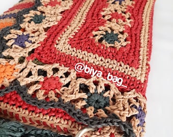 Puede incluir: Un bolso tejido a mano, colorido y con un diseño único. El bolso presenta un panel central rojo con motivos florales, bordeado por detalles beige, naranja y verde oscuro. El texto "@biya_bag_" es visible en el bolso.