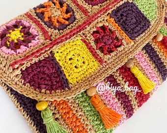 May include: A handmade, colorful crochet clutch with a flap closure. The bag features a patchwork design with squares in various colors, including pink, yellow, green, and purple. It has wooden bead accents and tassel details. The text "@biya bag" is visible.