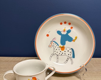 May include: A white ceramic bowl and mug set with a circus theme. The bowl has a blue and orange rim and features a clown riding a spotted horse. The mug has a clown illustration in orange and black.