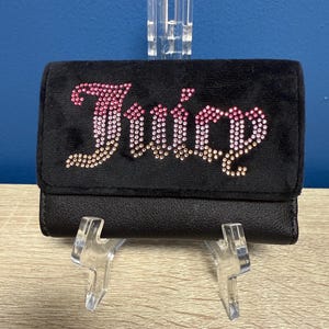 May include: Black wallet with the word "Juicy" spelled out in pink and gold rhinestones. The wallet is made of a soft, velvety material and has a fold-over flap. The wallet is displayed on a clear stand.