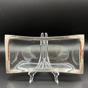 May include: A rectangular glass tray featuring a clear, textured center and a wide, polished silver-toned metallic border. The tray has slightly curved, concave sides and is displayed on a clear plastic stand for support.