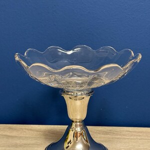 May include: A clear glass footed bowl with a scalloped rim. The bowl sits atop a silver-colored pedestal base. The base has a wide, round foot and a cylindrical stem. The background is a solid blue.