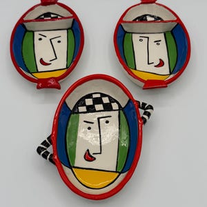 May include: Three ceramic dishes featuring hand-painted abstract faces. Two smaller round dishes have red rims and black and white checkered details. One larger oval dish has a red rim and black and white striped handles on the sides.