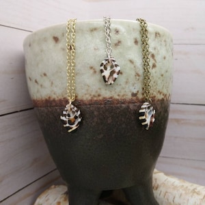 May include: Three gold necklaces with shell pendants hanging from a brown ceramic pot with a white glaze. The necklaces are on a light wood surface.
