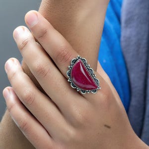 Peut inclure: Une bague en argent avec une grande pierre précieuse rouge foncé en forme de demi-lune sertie dans un cadre en argent décoratif.