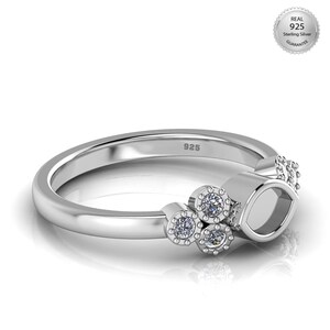 Könnte beinhalten: Ein silberner Ring mit poliertem Finish. Der Ring hat drei kleine Cluster aus klaren Edelsteinen und eine größere ovale Fassung. Das Band ist mit "925" gestempelt. Ein "Real 925 Sterling Silver Guarantee" Siegel ist sichtbar.