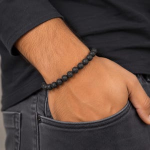 Può includere: Un braccialetto di perline composto da piccole sfere nere in pietra lavica con una superficie porosa e ruvida. Le perline sono infilate in un unico filo continuo, creando un design circolare uniforme che si adatta al polso.