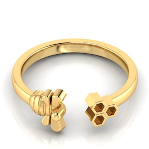 Puede incluir: Un anillo abierto de color dorado con un diseño de abeja y panal. La abeja está en un extremo y el panal en el otro. El anillo está hecho de un metal brillante y reflectante.