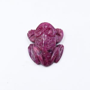Puede incluir: Una figurita de rana de rubí rosa tallada. La rana está sentada con las patas extendidas y tiene detalles elaborados.