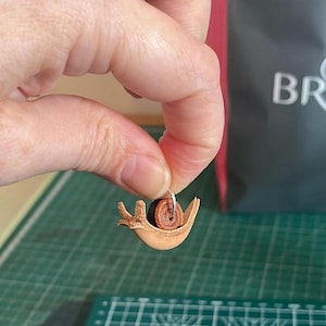 May include: A small, brown leather snail charm with a silver loop for hanging. The snail is curled up in its shell, which is made of brown leather.