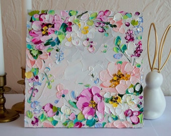 Op de afbeelding: Een vierkant canvas schilderij met een bloemenmotief. Het schilderij toont roze, witte en gele bloemen met groene bladeren op een lichtgrijze achtergrond.